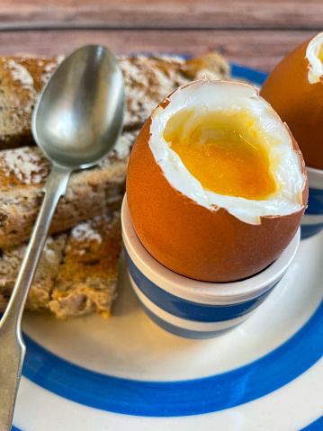 Stock photo showing elevated view of Cornish ware crockery of tea plate and egg cups with wholemeal toast soldiers and topped soft-boiled eggs showing runny yellow yolks.