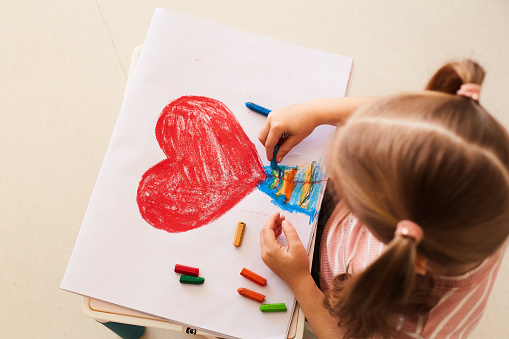 Horizontal photo. The girl draws a large red heart on a White leaf. There are colored pencils next to them.