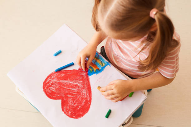 Horizontal photo. The girl draws a large red heart on a White leaf. There are colored pencils next to them.