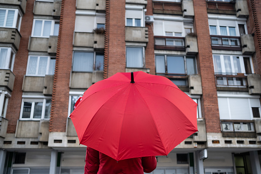 girl in a red jacket with a red umbrella stands in front of buildings on a cloudy day