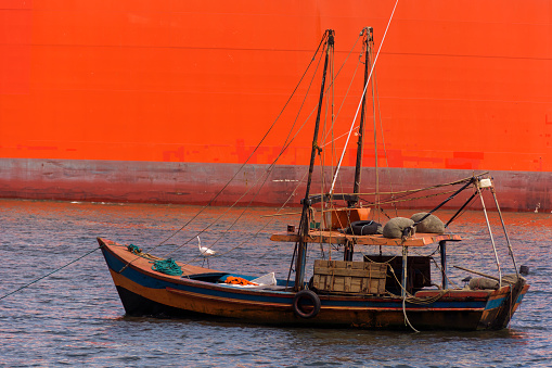 Colorful fishing boat stopped at sea with a white heron at the bow and the hull of a big red ship passing in the background.
