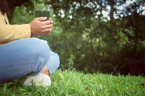 Relax legs of a woman sitting alone on green grass in the park with coffee or tea cup in hand. Person holding glass of water in left composition with copy space for inspirational text message.