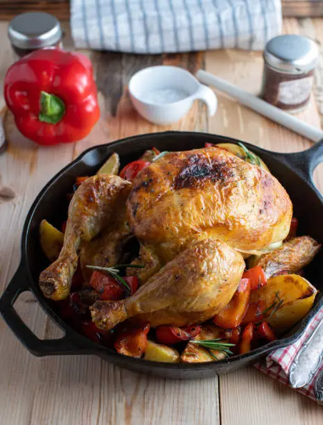 Homemade delicious oven dish with roast whole chicken, roasted potatoes and bell peppers. Served in a cast iron pan on wooden table background. Ready to eat