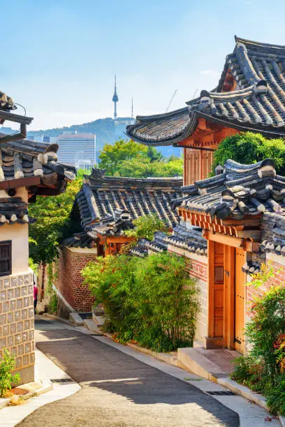 Cozy old narrow street and traditional Korean houses of Bukchon Hanok Village in Seoul, South Korea. Seoul Tower on Namsan Mountain is visible on blue sky background. Beautiful cityscape on sunny day.