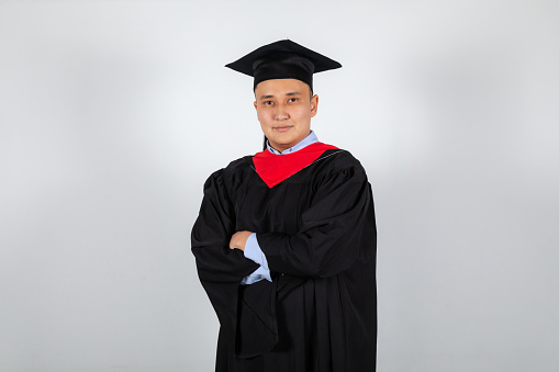 Portrait of graduate male student in mortarboard and bachelor gown isolated on white background crossed hands
