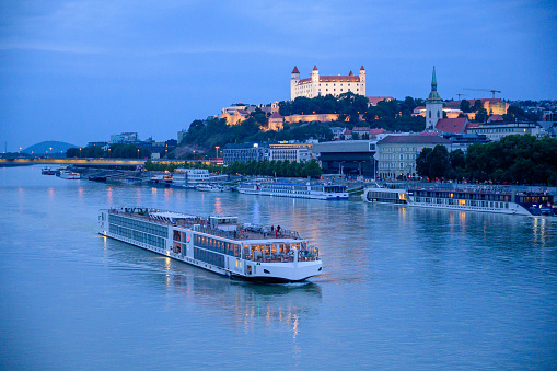 Bratislava, Slovakia - July 9, 2019: A river cruise boat passes along the River Danube in front of Bratislava Castle in the blue light of evening