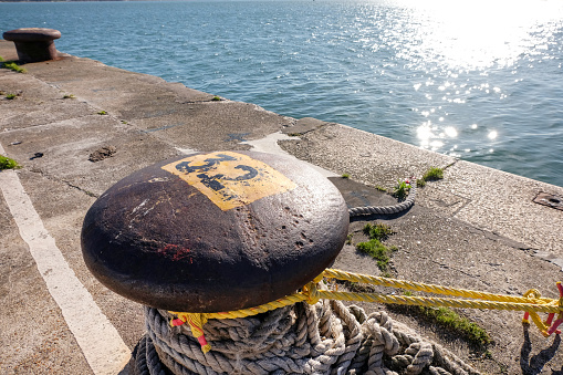 Huge metal bollard with different sized ropes on a quay wall on the shore and sun reflections in the water, copy space