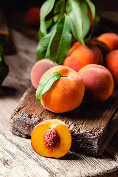 Peaches with leaves on dark wooden board with peach in halves with peach seed stone. Composition with ripe juicy peaches Harvest for food. Fresh organic fruit.
