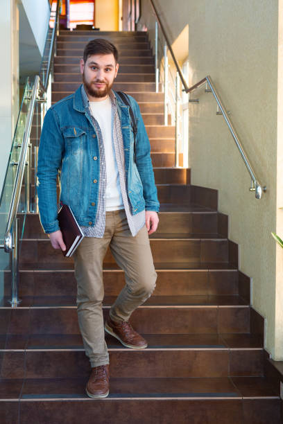 istock Student on brown stairs. Caucasian smiling handsome bearded man with backpack notebooks in reopen university campus hallway. Freelancer in modern coworking office 1369979890