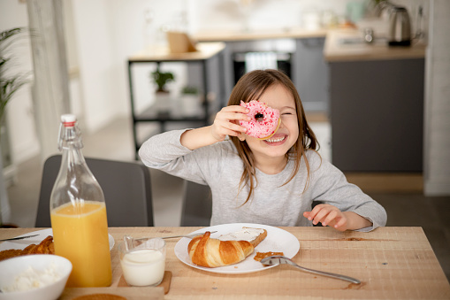 Portrait of a little cheerful Caucasian girl enjoying a sweet meal at home, having a doughnut and looking through it, playing with food
