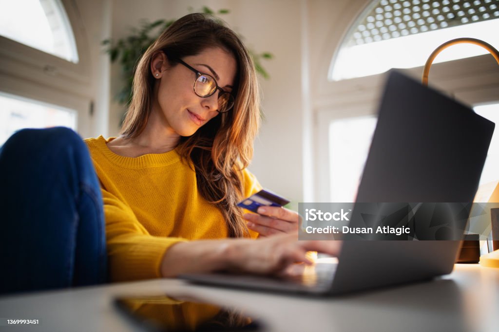Beautiful young woman working at home A beautiful young woman enjoying working at home on her laptop in cozy and bright apartment wearing yellow sweater and shopping online paying with credit card Credit Card Stock Photo