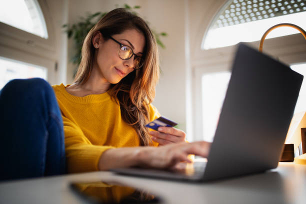 Beautiful young woman working at home A beautiful young woman enjoying working at home on her laptop in cozy and bright apartment wearing yellow sweater and shopping online paying with credit card home finances photos stock pictures, royalty-free photos & images