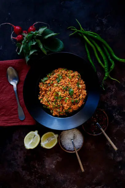 Istanbul, Turkey-February 11, 2022: Kısır on black large deep plate. Around the dinner plate are small bunches of red radish, green chili peppers, sliced lemon, sea salt and cayenne pepper spice balls. Kisir is a traditional dish made with bulgur, tomato paste, hot and sour spices and fresh greens. Kisir is more of a salad variety. There is also a silver spoon on a coral linen napkin. The plate is on a rough black-brown surface with a rust effect. The plate is bright and the surrounding of the plate is darker. Shot with Canon EOS R5.