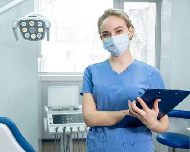 Dentist's assistant or female dentist in protective face mask on the background of dentistry room Dentist's assistant or female dentist in protective face mask on the background of dentistry room. Portrait of young female doctor wearing a blue uniform. Pretty nurse is looking at camera with smile dental hygienist stock pictures, royalty-free photos & images