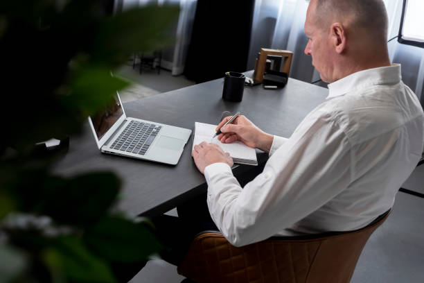 istock Mature man sitting at table with laptop, taking notes in notebook. Businessman n formal clothes working or studying in office. Online education or work on computer concept. 1369998425