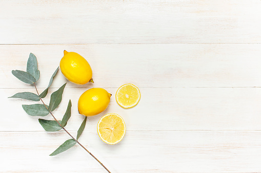 Whole and sliced fresh lemon, eucalyptus leaves on white wooden background. Flat lay, top view, copy space. Minimal fruit concept design. Yellow citrus lemons