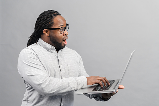 Black man standing with laptop on grey background, typing, concept of humans emotions, feeling stunned by online news looking at computer screen, trader investor surprised by stock market changes