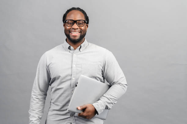 Working with joy concept Working with joy. Smiling cheerful african american business man in glasses wearing classic shirt, standing with laptop, pc computer, looking at camera, isolated on grey background studio portrait black man stock pictures, royalty-free photos & images