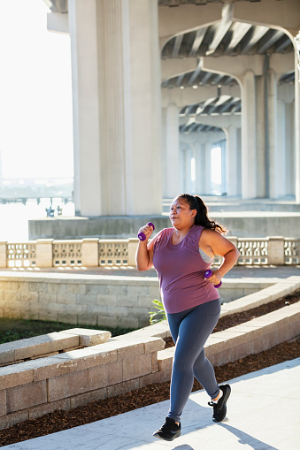 A Pacific Islander woman running on a city waterfront, under a bridge. Large concrete columns are in the background.