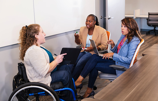 A multiracial group of three businesswomen having a meeting in an office board room. The two African-American women and their coworker, in a wheelchair, are looking at one another, talking, and pointing with serious expressions.