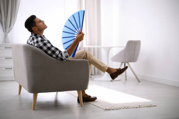 istock Man with hand fan sitting on sofa. Summer season 1370016362