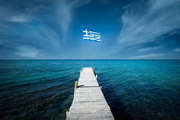 Photo of Greek flag on a beautiful sunny day at the end of a white wooden pier. Corfu Greece