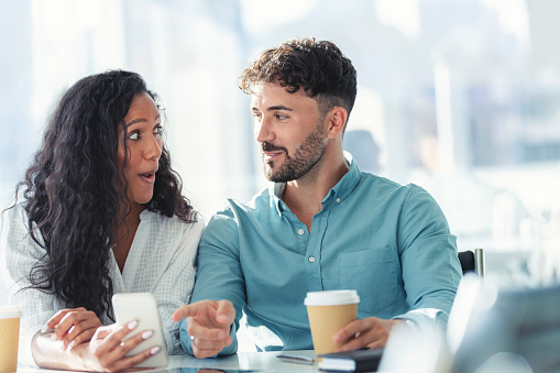 Businessman and businesswoman looking at a mobile phone. Woman has a shocked look on her face. Multi ethnic group with African American and Caucasian. They are casually dressed and having a discussion. They are drinking coffee. Could be on a video call, online shopping or sending a text message.