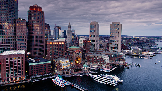 Aerial shot of Downtown Boston, Massachusetts at sunset on an overcast evening, taken from over the Harbor and looking towards office towers, Rowe's Wharf ferry terminal and other buildings on the waterfront. \n\nAuthorization was obtained from the FAA for this operation in restricted airspace.