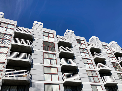 Exterior view of typical multifamily mid-rise residential apartment building with balconies and identical windows blinds under blue sky.