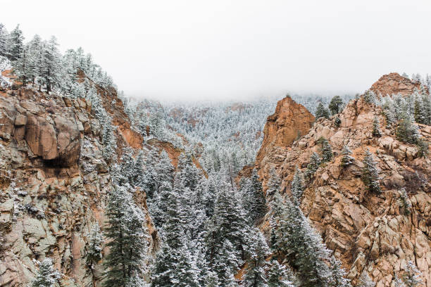 A light snow storm passes through a mountain ridge in central Colorado. The orange rock of the sharply ridged mountainside meets the forest green pines lightly covered in snow during an overcast March day.