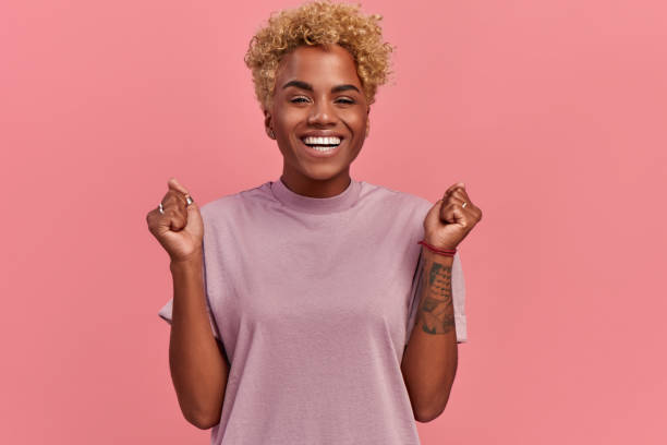 Portrait of overjoyed black female clenches fists with happiness, opens mouth widely as shouts loudly, celebrates her success, poses against pink background. Happy African woman with joyful expression Portrait of overjoyed black female clenches fists with happiness, opens mouth widely as shouts loudly, celebrates her success, poses against pink background. Happy African woman with joyful expression clenching teeth stock pictures, royalty-free photos & images