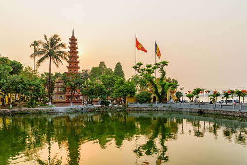 Hanoi, Vietnam - April 19, 2019: Wonderful sunset view of the Tran Quoc Pagoda and West Lake. The flag of Vietnam (red flag with a gold star) and the Vietnamese Buddhist flag fluttering over the lake.