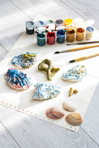 Child's craft place with shells have been painted different colors, paintbrushes and gouache paints. Child painting seashells for DIY project.