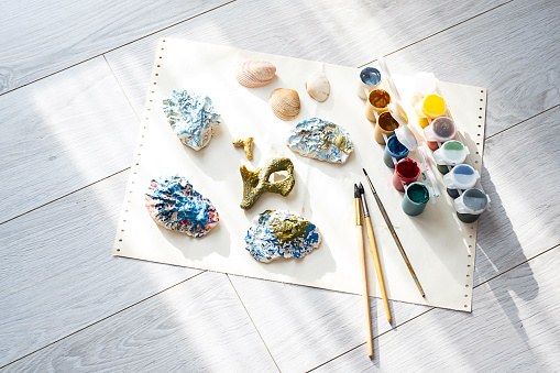 Child's craft place with shells have been painted different colors, paintbrushes and gouache paints. Child painting seashells for DIY project.
