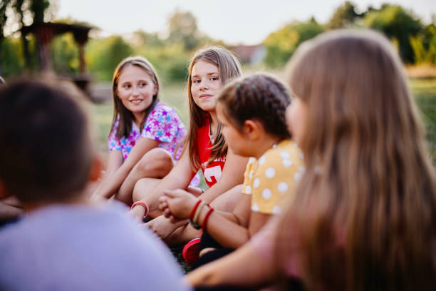 Group of kids bulling girl at park during summer camp - Little girl looking camera, sitting lonely while friends playing and having fun.