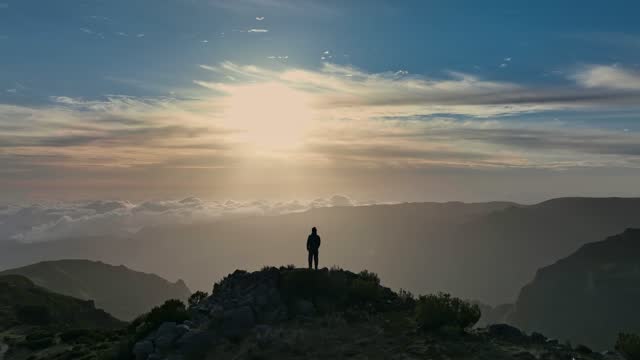 Climber in the mountains achieved his goal and enjoys wonderful nature. Aerial view of magnificent sunrise in the mountains of Madeira island. Man stands on top of mountain and looks at the sunrise sky.