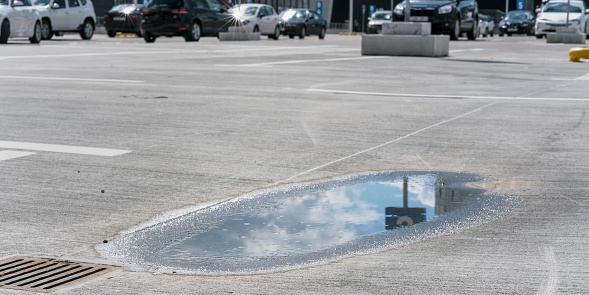 large rainy puddle reflects blue sky with white clouds and road sign on asphalt parking lot against distant vehicles