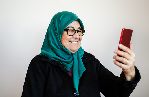 Senior muslim woman wearing a green headscarf taking a selfie, using smartphone at home, Day light
