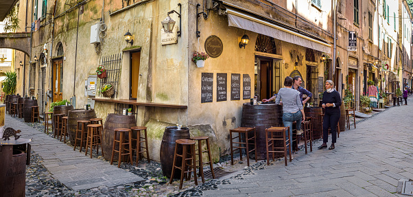 Finalborgo, Finale Ligure, Italy. May 5, 2021. View of a typical trattoria wine bar restaurant in Via Giovanni Nicotera with people talking near the outside wooden barrel-shaped tables.