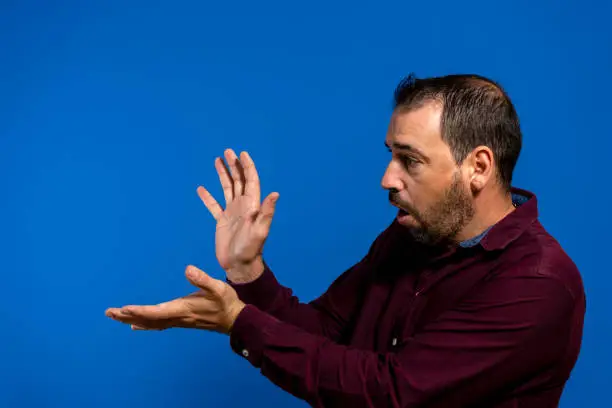 Latin bearded man dressed in a purple shirt isolated on blue studio background, he is throwing a kame hame imitating Son Goku from Dragon Ball, one of his youth heroes.