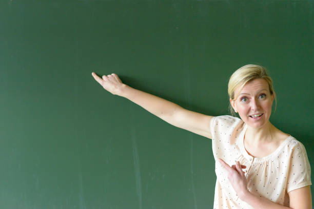 Female teacher pointing at copy space place on chalkboard, looking at camera and smiling