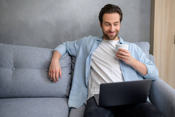 istock Optimistic man sitting on the couch and using laptop computer. Happy businessman working remotely in relaxed pose, drinking coffee 1370106124
