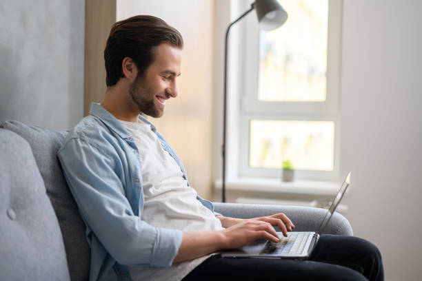 istock Optimistic man sitting on the couch and using laptop computer for messaging. Happy businessman working remotely in relaxed pose 1370106153