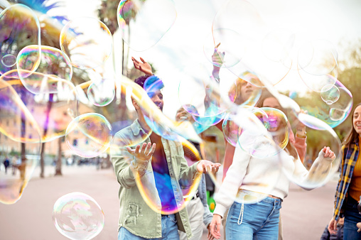 Group of happy friends having fun with soap bubbles in the town square - Millennial guys and girls having fun around the streets - University students on travel vacations