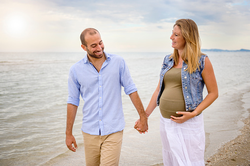 Happy smiling man holding his pregnant girlfriend's hand and look at her belly - Young loving couple walking on the beach on a sunny day - Multiethnic lovers expecting a baby