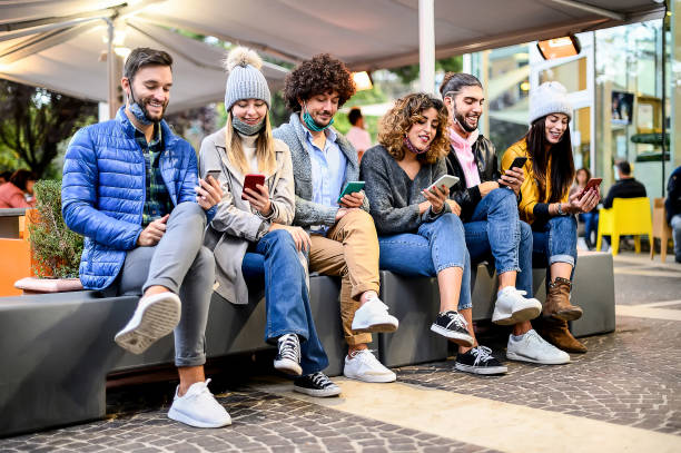 istock Young happy friends with face mask down sitting on bench using smartphone watching social media content online outside the bar - Concept of youth, tech and friendship - People Working Together 1370110067