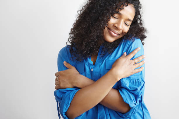 Self love concept. Isolated shot of beautiful pleased young dark skinned female with voluminous hairstyle embracing herself and closing eyes, enjoying soft fabric of her new stylish blue shirt Self love concept. Isolated shot of beautiful pleased young dark skinned female with voluminous hairstyle embracing herself and closing eyes, enjoying soft fabric of her new stylish blue shirt hugging self stock pictures, royalty-free photos & images