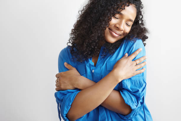 self love concept. isolated shot of beautiful pleased young dark skinned female with voluminous hairstyle embracing herself and closing eyes, enjoying soft fabric of her new stylish blue shirt - self love imagens e fotografias de stock
