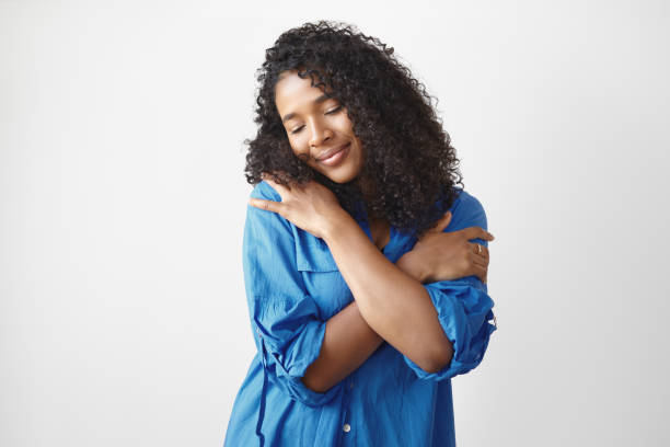 Studio shot of gorgeous charming young Afro American lady in stylish blue shirt having pleased happy facial expression, closing eyes and smiling, embracing herself, full with love and appreciation Studio shot of gorgeous charming young Afro American lady in stylish blue shirt having pleased happy facial expression, closing eyes and smiling, embracing herself, full with love and appreciation alternative pose photos stock pictures, royalty-free photos & images