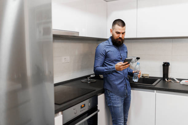 istock Portrait of young man using his smart phone and drinking water in his kitchen 1370115852